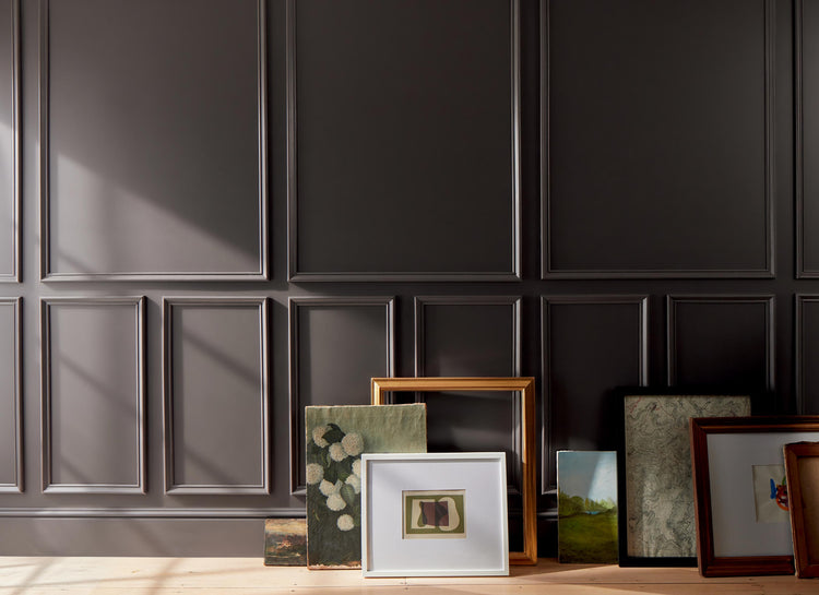 Artwork and frames arranged on a floor against a deep chocolate brown-painted wall with decorative molding.